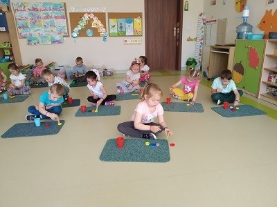 Dzieci siedzą przed dywanikiem, na nim mają kolorowe nakrętki i kubki 